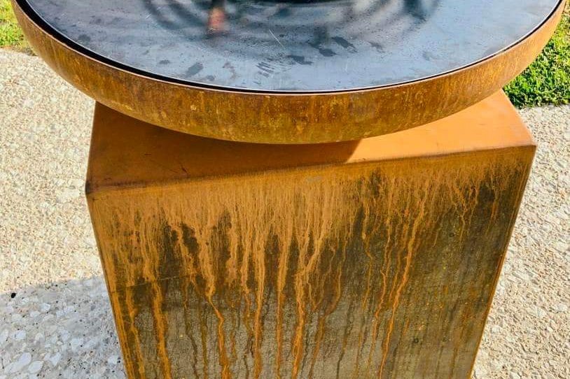 Braséro rond en acier corten sur un meuble corten, posé sur une allée gravillonnée