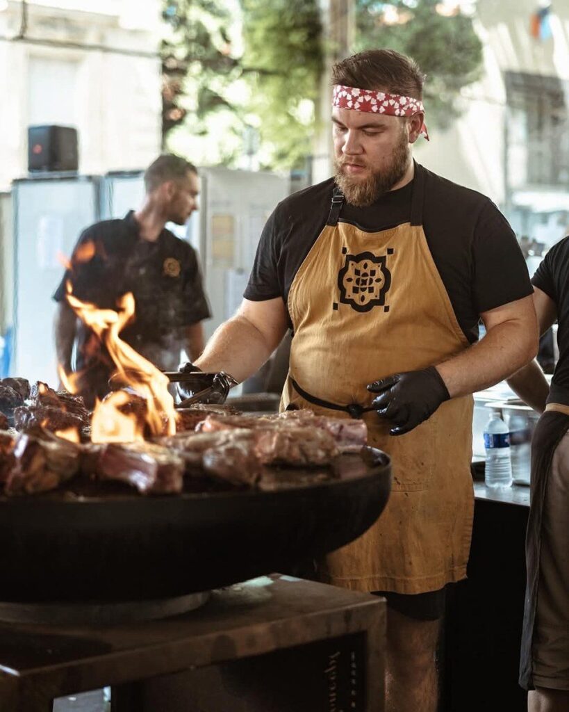 Cuisinier au bandana devant un braséro de 100 chargé de viandes