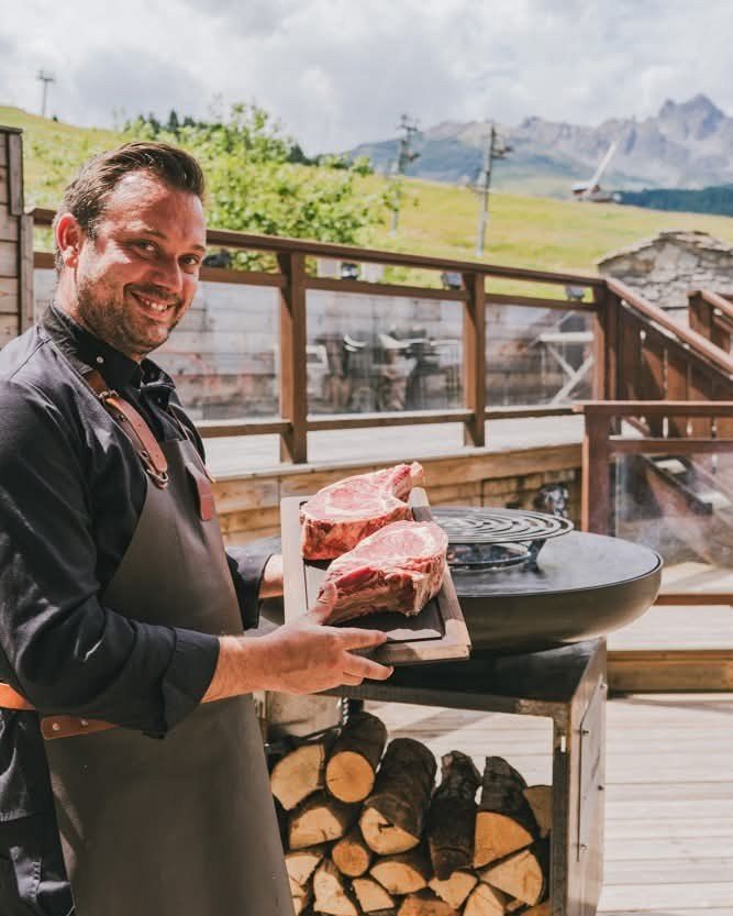 Cuisinier souriant présentant deux côtes de bœuf au-dessus du braséro