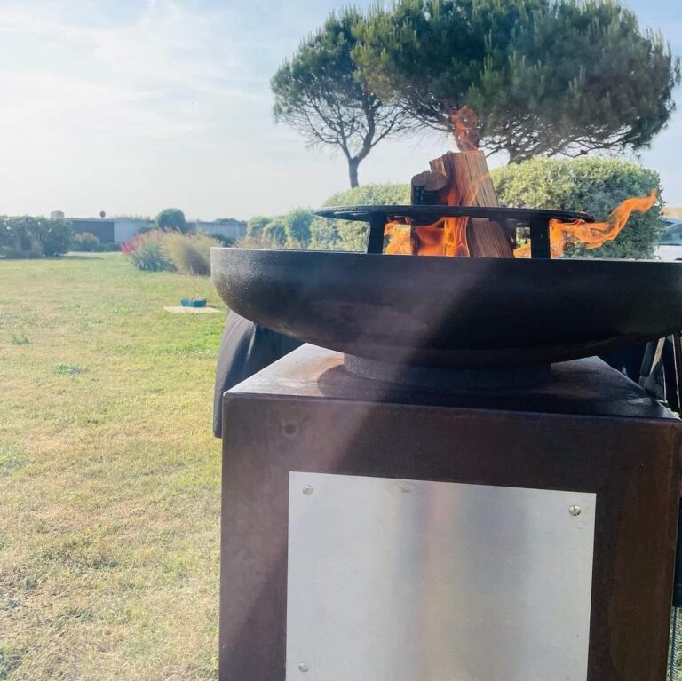 Braséro rond de 80 sur meuble en corten, flammes et pins au bord d'un jardin