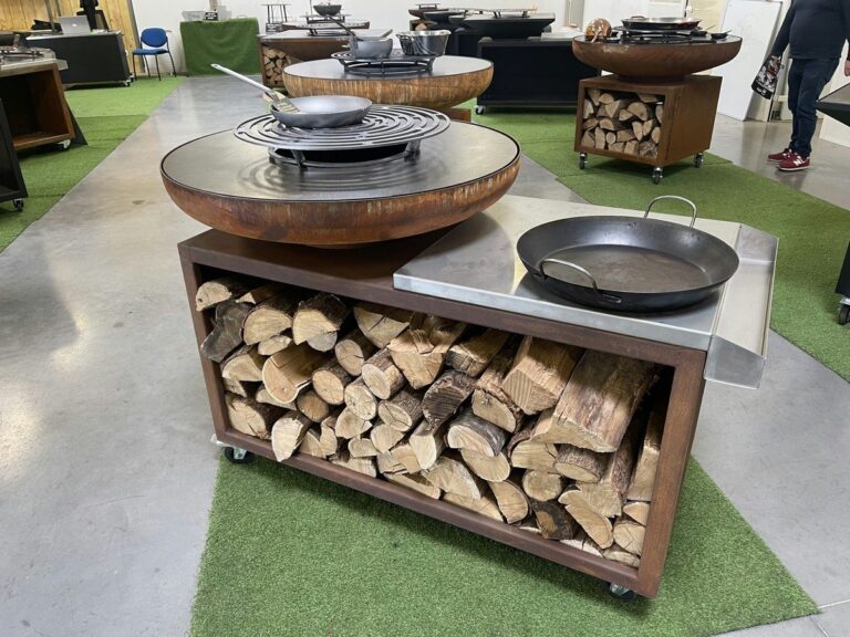 Braséro rond en corten sur meuble ouvert rempli de bûches, poêle posée à côté