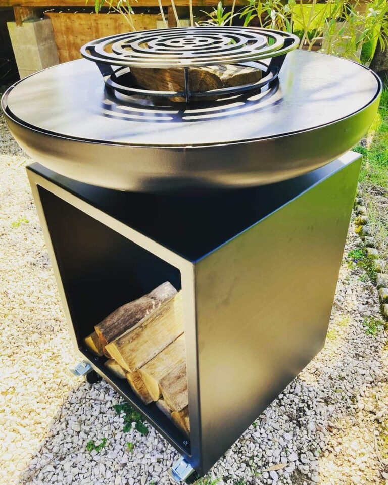 Braséro rond noir sur meuble avec réserve de bûches, posé sur du gravier