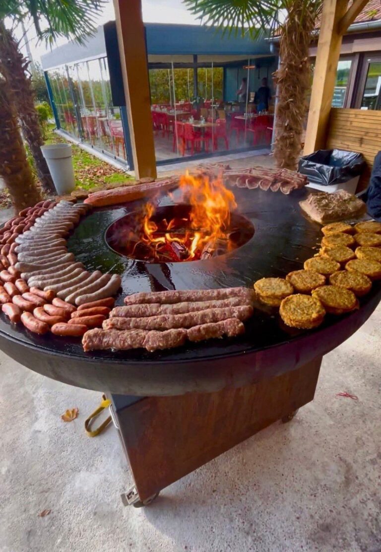 Braséro rond sur meuble en corten garni de saucisses, merguez et galettes autour du foyer, devant un restaurant