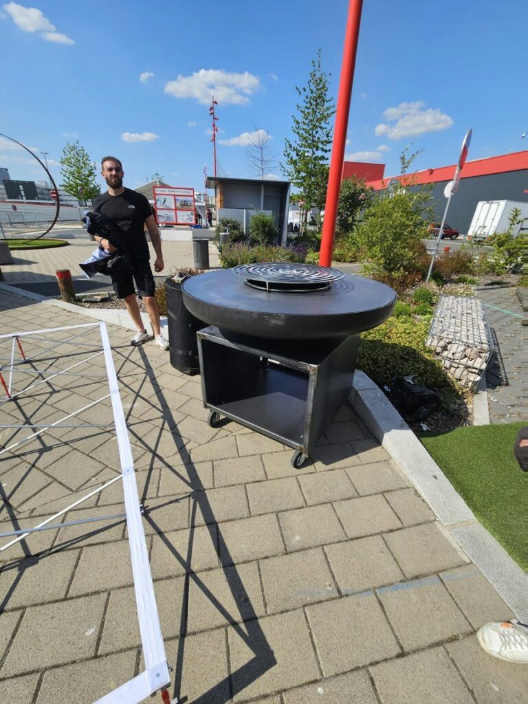 Braséro rond sur meuble noir ouvert au bord d'une allée pavée, cuisinier au fond