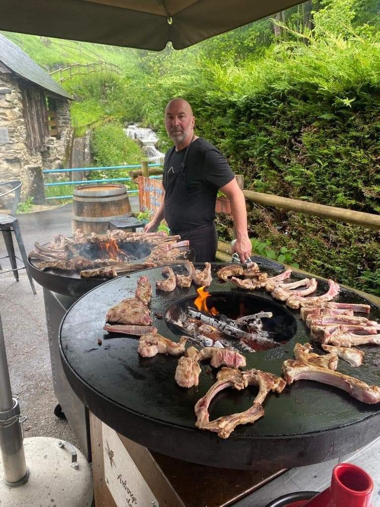 Cuisinier devant un braséro rond garni de côtes d'agneau et d'un second foyer au fond, terrasse d'auberge en montagne
