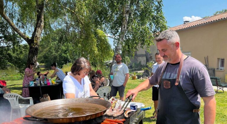 Cuisinier en tablier de cuir devant une plancha de saucisses et une paella géante, au jardin