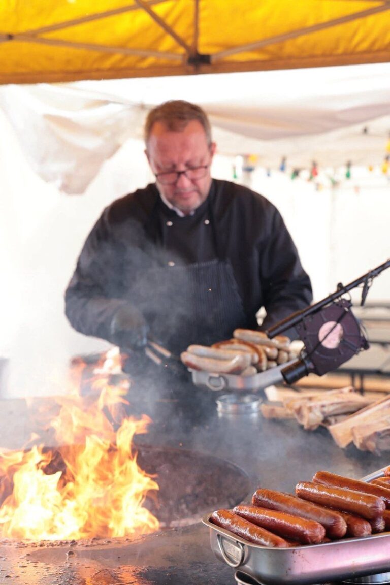 Cuisinier en veste noire devant un braséro en flammes garni de saucisses, sous une tente jaune