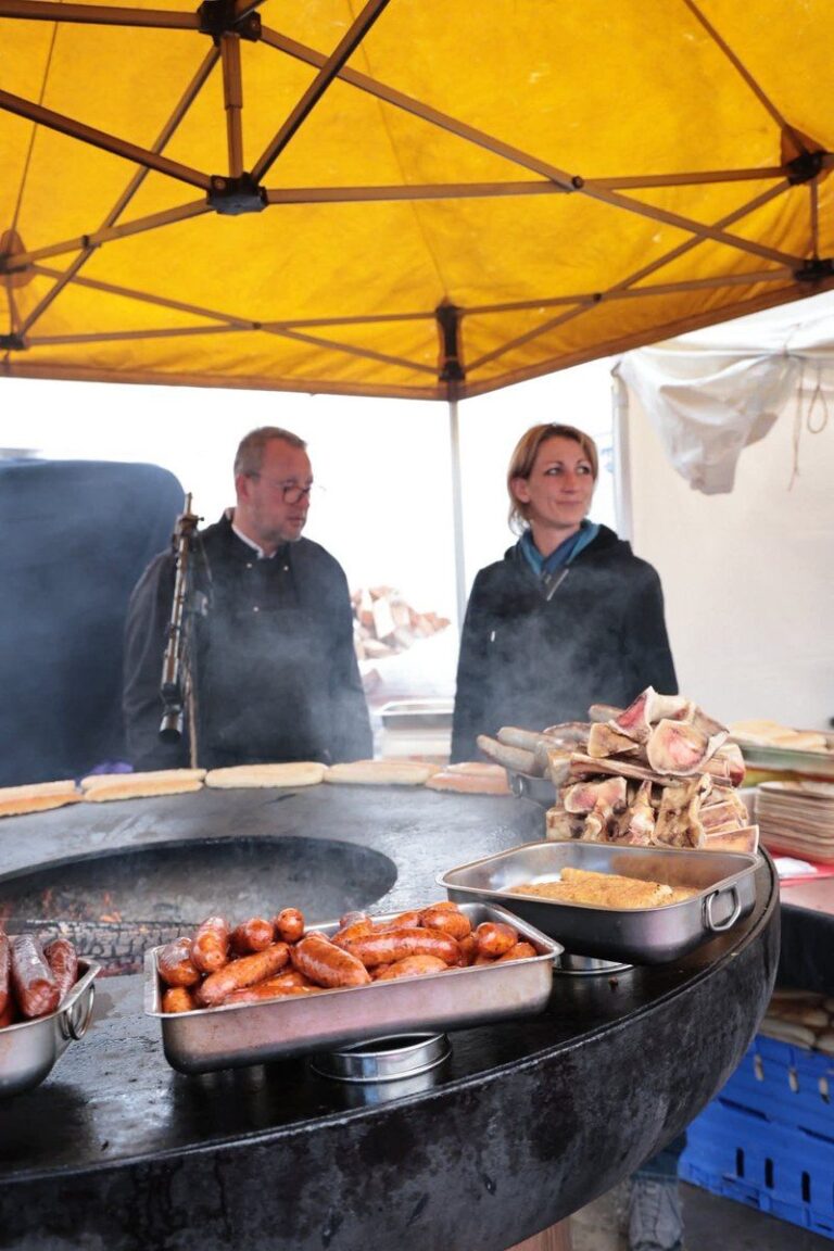 Deux cuisiniers sous une tente jaune devant un braséro garni de saucisses, d'os à moelle et de bacs