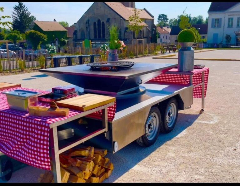 Remorque braséro à double essieu avec ses tables à nappe vichy, installée sur un parvis