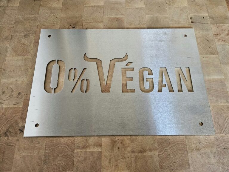 Plaque inox au logo 0% Végan découpé, avec quatre trous de fixation, posée sur un billot de boucher