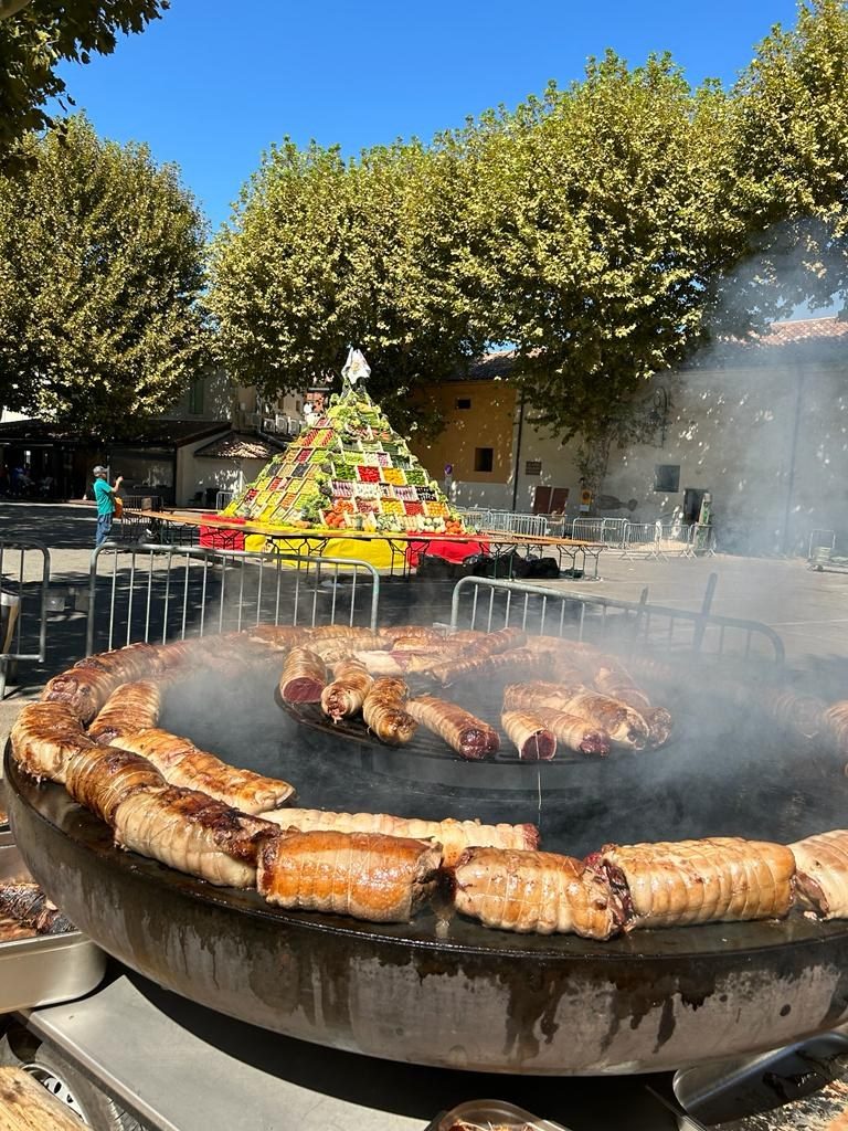 Très grand braséro rond garni de grillades sur une place de village, pyramide de légumes au fond