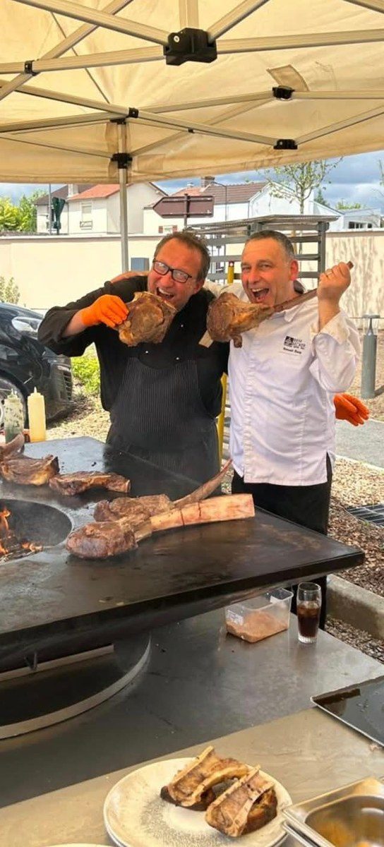 Deux cuisiniers présentant des pièces de bœuf devant la plancha d'une remorque braséro