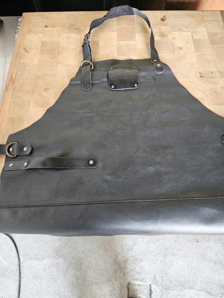 Tablier en cuir noir avec sa plaque 0% Végan, posé à plat sur un billot de boucher